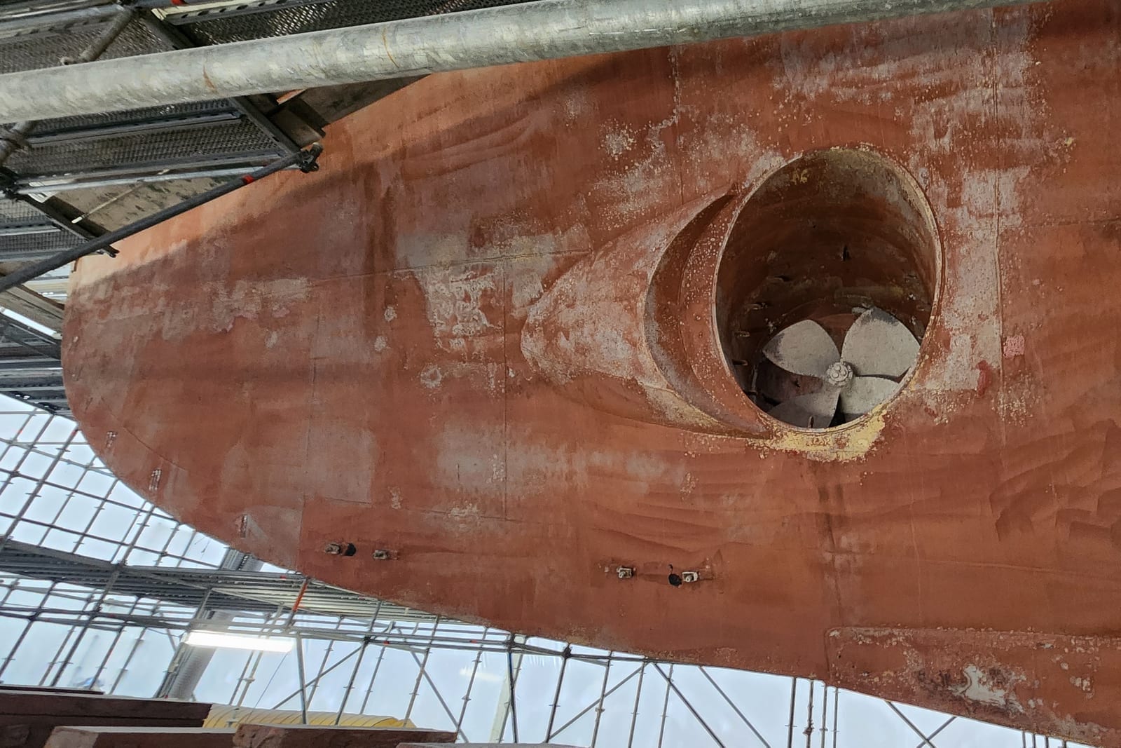 Yacht hull under scaffolding with the bow-thruster tunnel exposed during yard work.